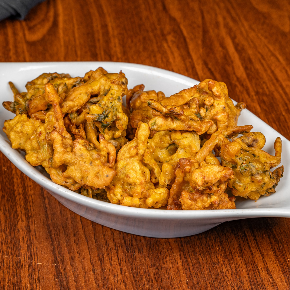 A white dish filled with golden-brown, crispy pakoras, a popular halal appetizer, on a wooden table.