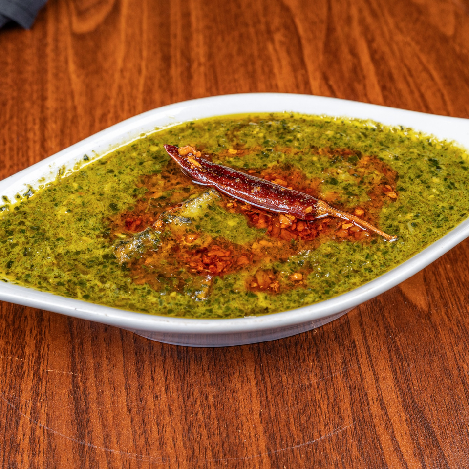 A white bowl of green curry with red chili flakes and a whole dried red chili on a wooden table.