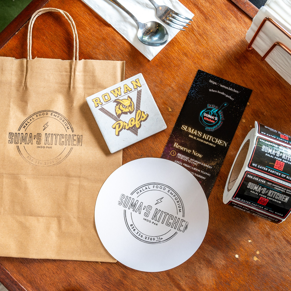 Suma's Kitchen branded paper bag, coaster, menu, and sticker roll on a wooden table.